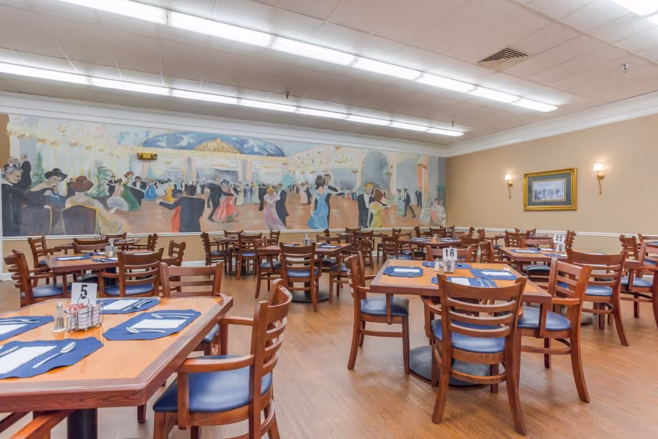 A dining room with multiple wooden tables and chairs arranged neatly. Each table has blue placemats, silverware, napkins, and condiment holders. The walls are beige with a large mural depicting people dancing in formal attire on one side and a framed picture on another wall. The room is well-lit with ceiling lights and wall sconces.