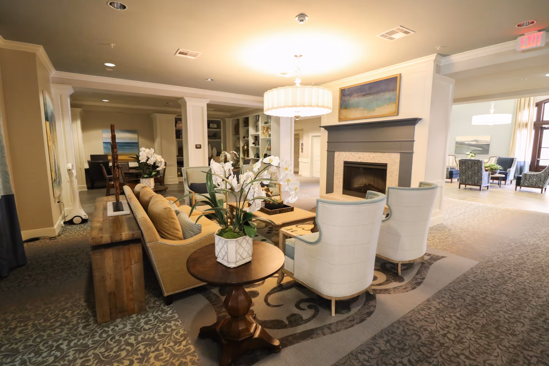 A spacious and well-lit living room area in an assisted living facility featuring a beige sofa, two white armchairs, a wooden side table with a white orchid plant, a fireplace with a painting above it, and additional seating areas visible in the background.