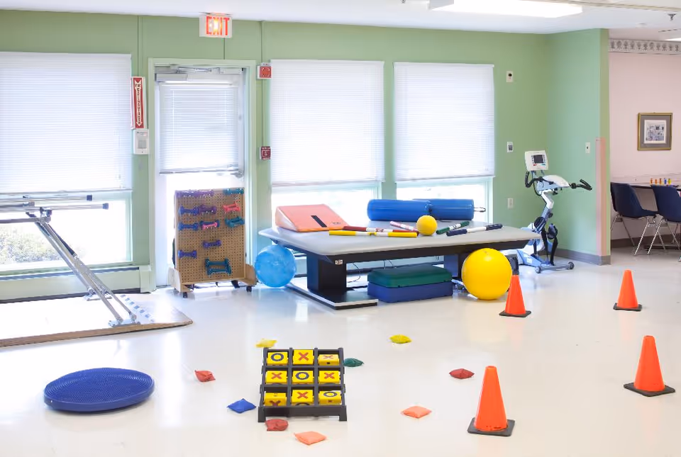Bright rehabilitation/activity room with a therapy table, exercise balls, cones, and various therapy equipment.