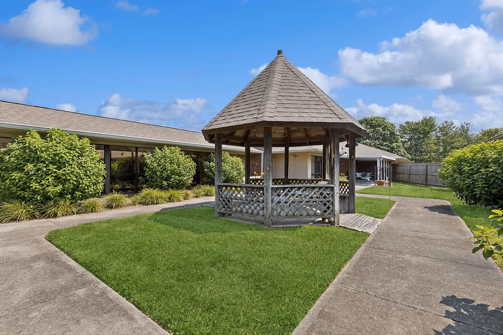 Outdoor courtyard area with a wooden gazebo in the center surrounded by green grass, concrete walkways, bushes, and a single-story building under a blue sky with some clouds.