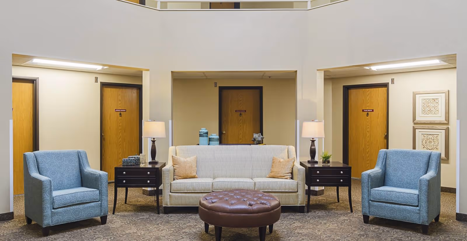 A cozy seating area in a senior living facility with a beige striped sofa flanked by two blue armchairs. There are two dark wooden side tables with lamps on either side of the sofa. Behind the seating area are three wooden doors and some decorative items on the walls and tables.
