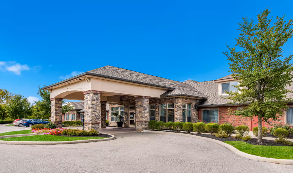 Exterior view of Dublin Glenn Memory Care facility showing a covered entrance with stone pillars, a curved driveway, landscaped bushes, a tree, and parked cars under a clear blue sky.