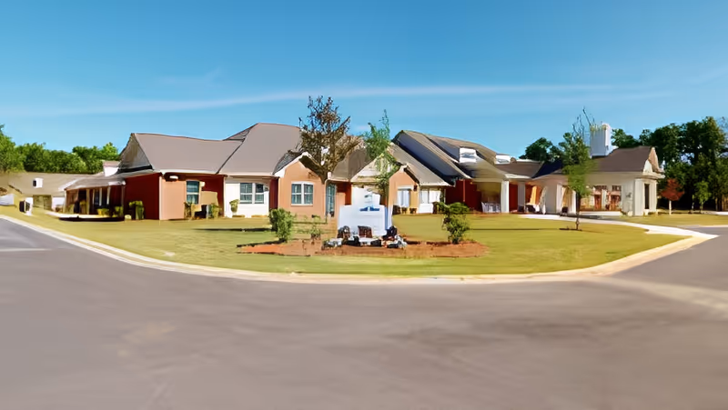 Single-story senior living building with a landscaped front lawn, circular driveway, and an entrance sign under a clear blue sky.