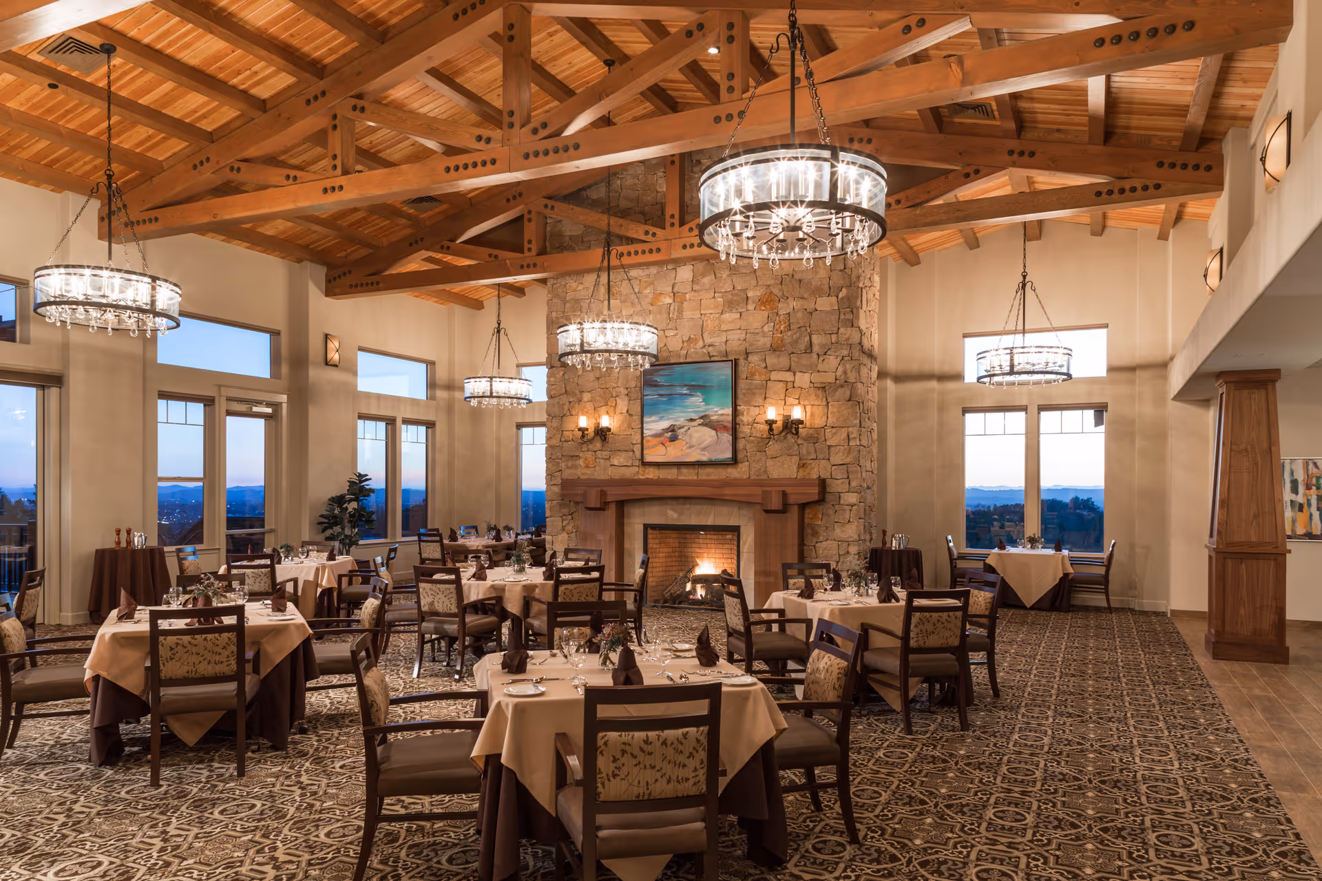 Elegant dining room with multiple tables covered in beige and brown tablecloths, set with glassware and napkins. The room features a high wooden beamed ceiling, large windows with scenic views, a stone fireplace with a painting above it, and several hanging chandeliers providing warm lighting.