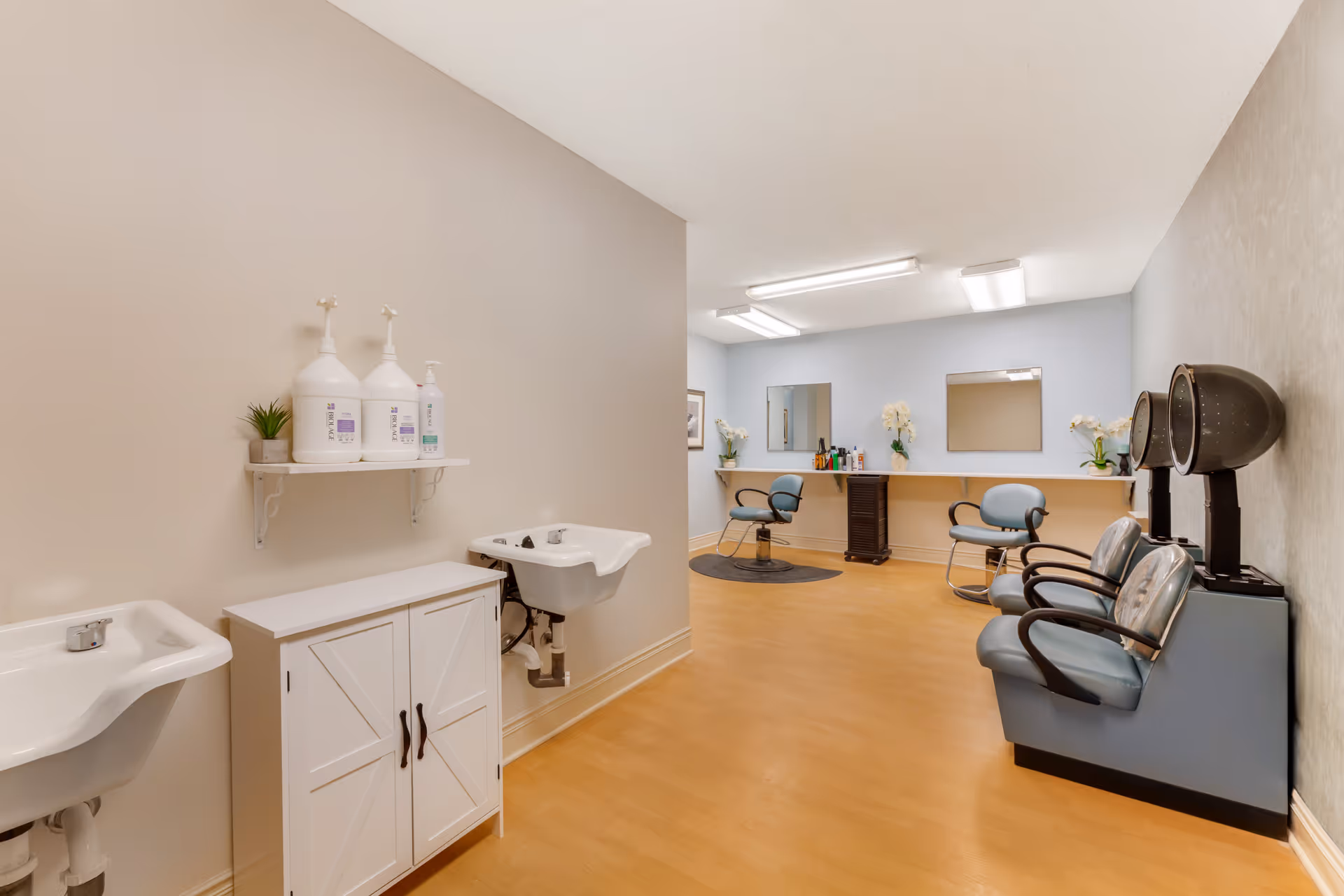 Interior view of a hair salon area in a senior living facility. The room features two hair washing sinks on the left wall with large bottles of hair care products on a shelf above a white cabinet. On the right side, there are two hair drying chairs with hooded dryers. Further into the room, there are two styling chairs in front of mirrors mounted on a light blue wall, with various hair products on a counter beneath the mirrors. The floor is a light wood laminate, and the room is brightly lit with ceiling lights.
