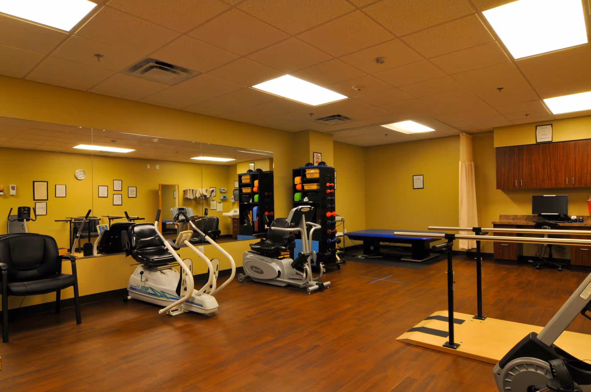 A fitness and rehabilitation room with exercise equipment including stationary bikes, a rack of colorful dumbbells, a padded therapy table, parallel bars for walking exercises, and a computer workstation. The room has wood flooring, yellow walls, and a large mirror on one side.