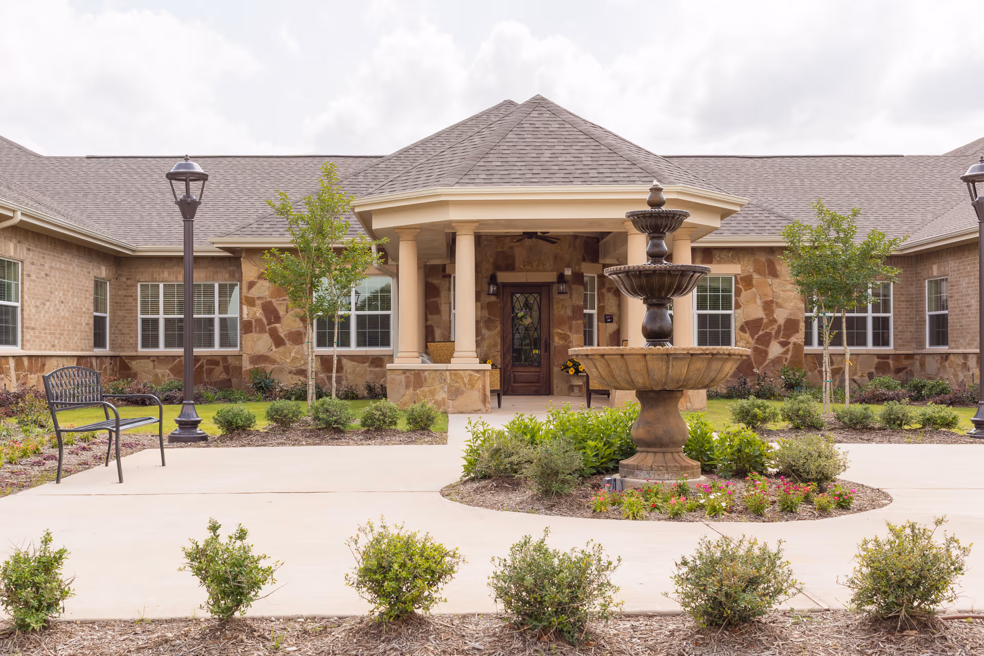 Front exterior view of a senior living facility with a stone and brick facade, a covered entrance supported by columns, a decorative fountain in the center of a landscaped circular garden, benches, and lamp posts along the walkway.