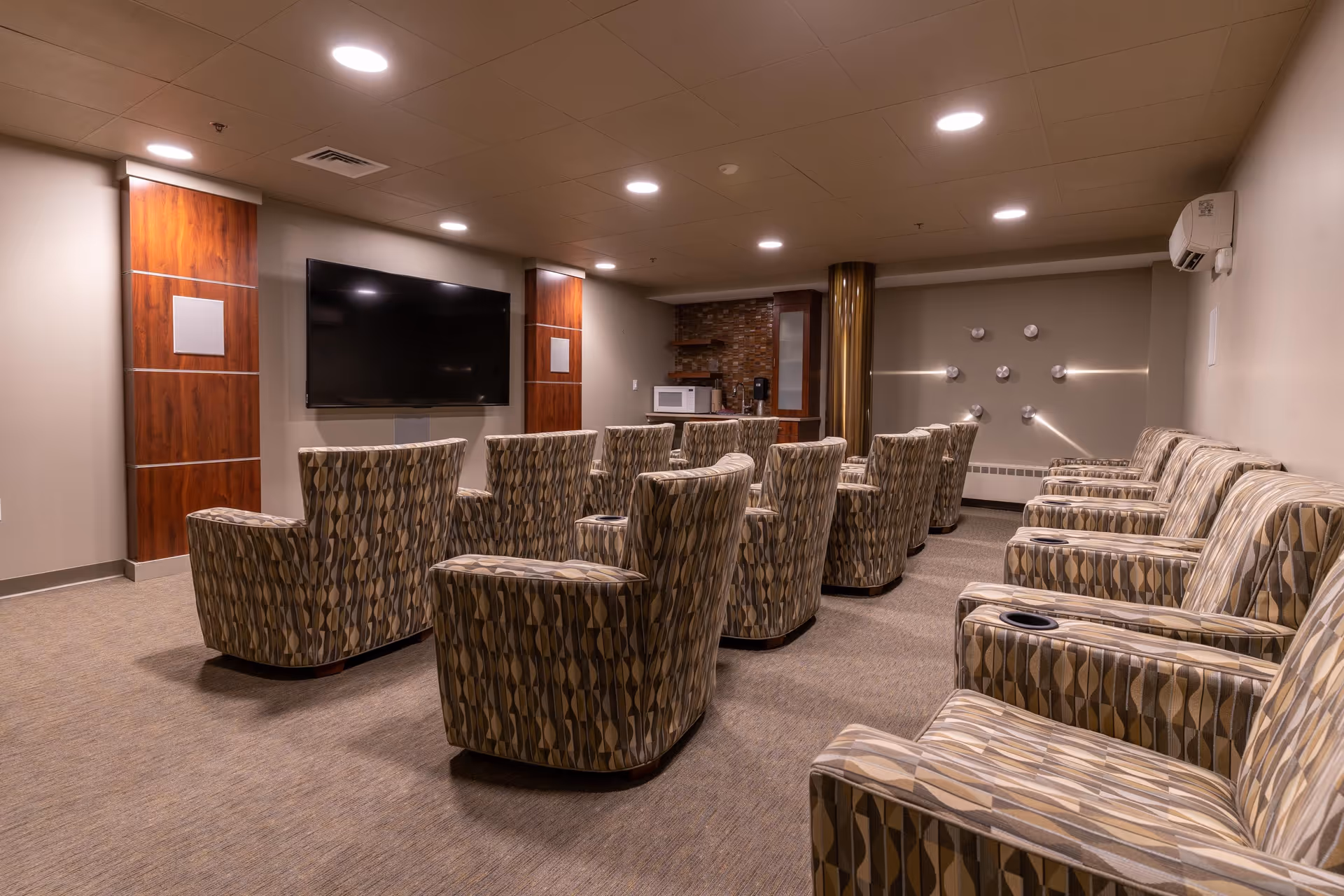 A cozy media room with multiple patterned armchairs arranged in rows facing a large flat-screen TV mounted on the wall. The room has a carpeted floor, recessed ceiling lights, and a small kitchenette area with a microwave and coffee maker in the back corner.