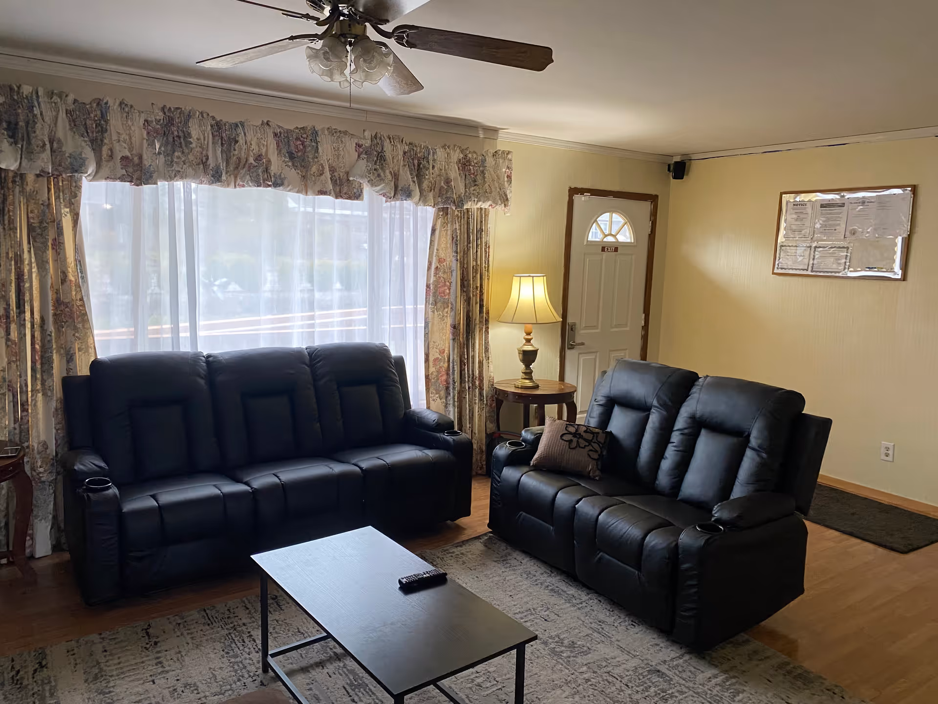 Living room with two black leather reclining sofas facing a coffee table, a large window with floral curtains, a side table lamp, and an entry door.