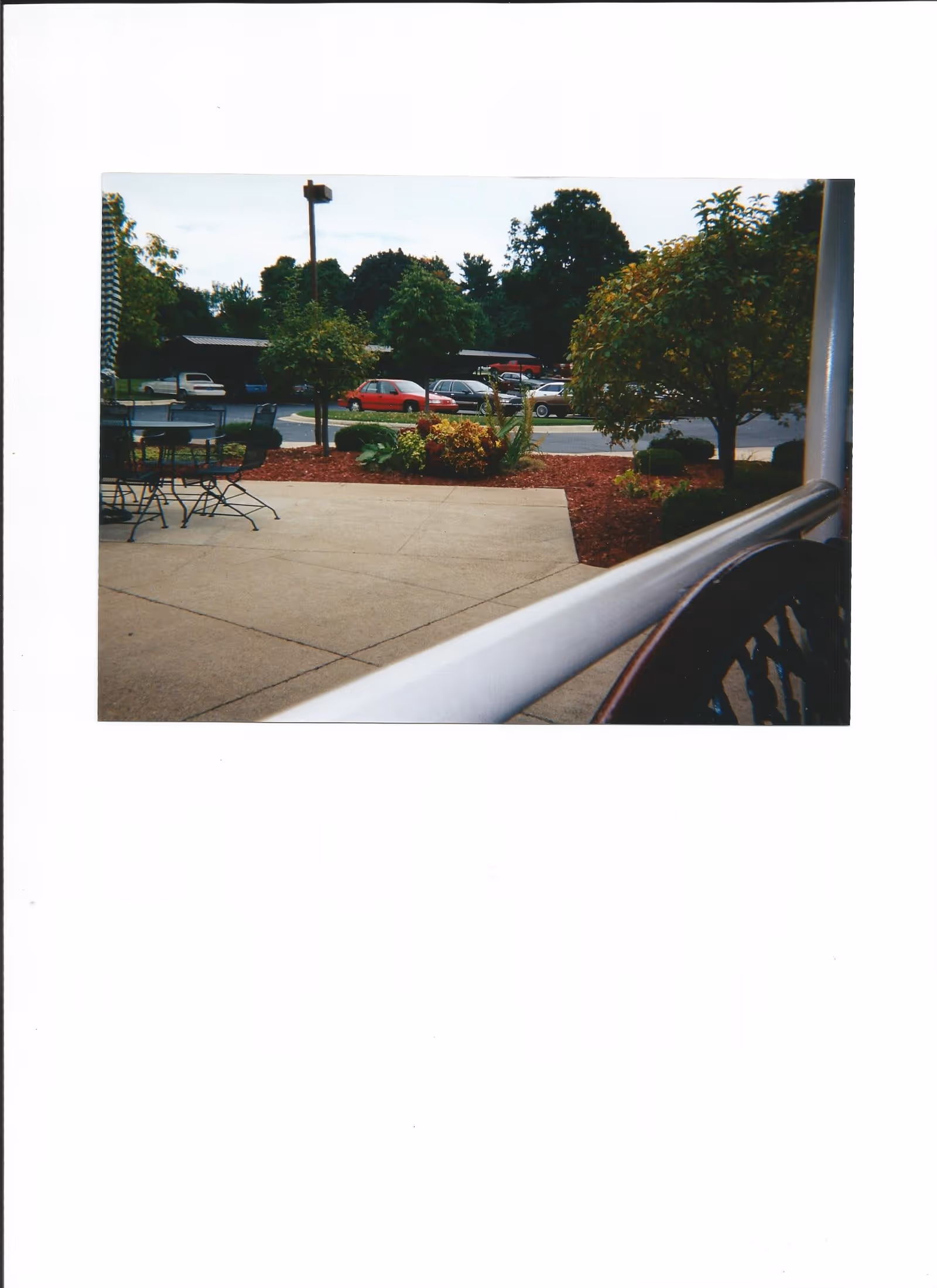 Patio area with metal tables and chairs, landscaped beds, a railing in the foreground, and parked cars and trees beyond.