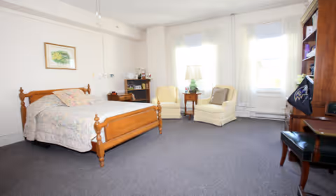 Bright, spacious assisted-living bedroom with a wooden bed, two armchairs by large windows, a lamp and a dresser.