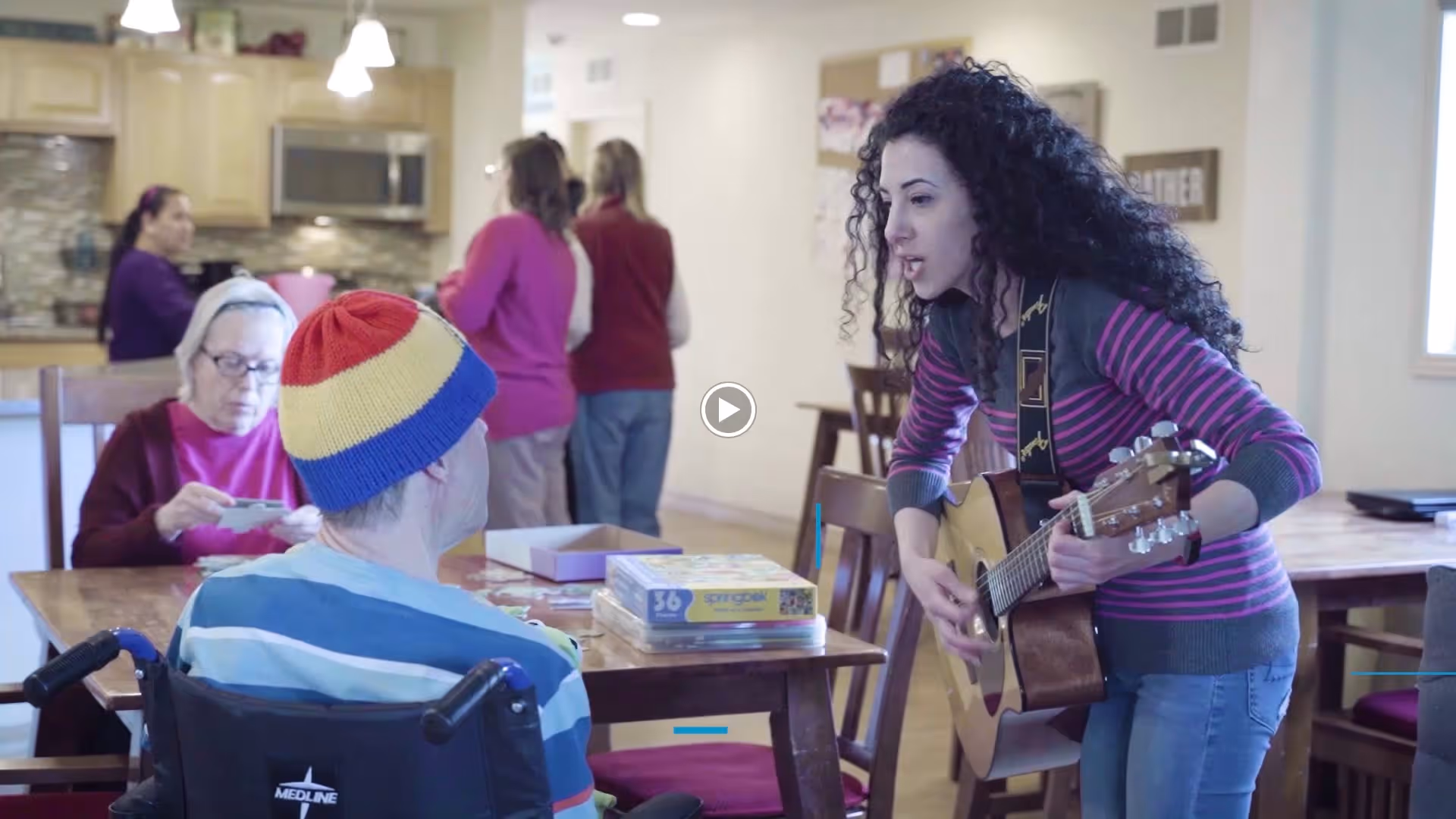 A woman with curly hair playing guitar and singing to an elderly man in a wheelchair wearing a colorful beanie, while other elderly people and staff interact in a kitchen and dining area in the background.