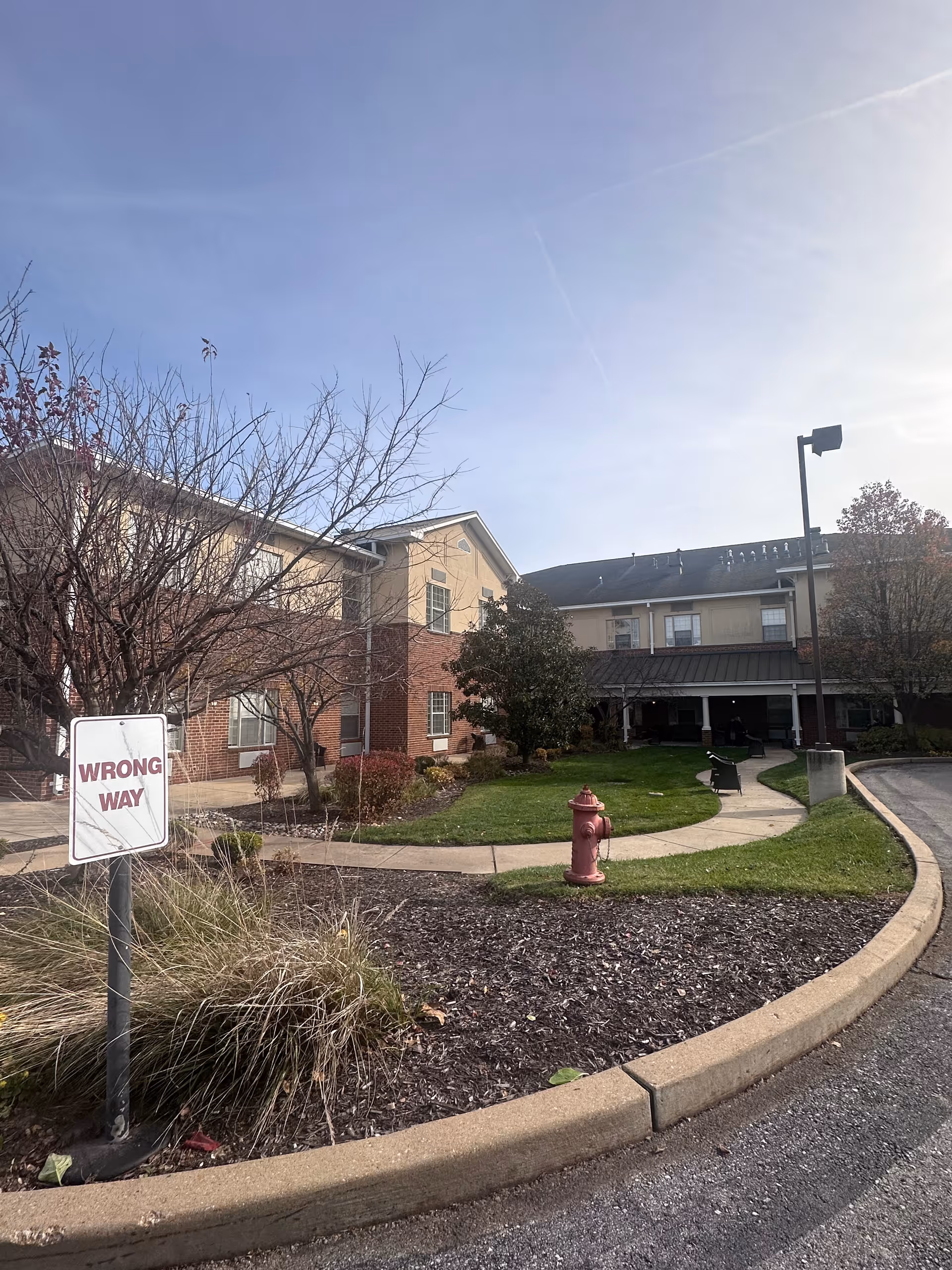 Exterior view of a senior living facility building with a curved sidewalk, green lawn, a red fire hydrant, leafless trees, and a 'WRONG WAY' sign near the curb under a clear sky.