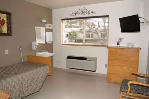 A furnished senior living bedroom with bed, dresser, wall-mounted TV, window, and chair.