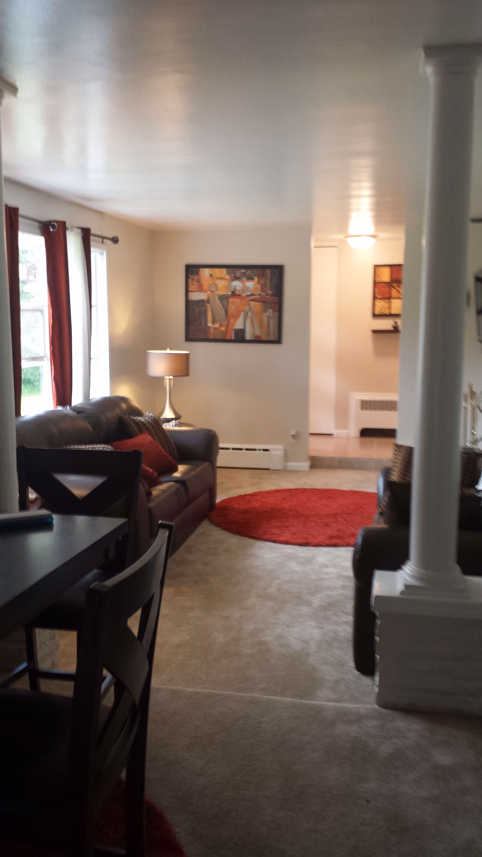 Interior view of a living room with beige carpet, a dark brown leather sofa with red and brown cushions, a round red rug, a table lamp on a side table, and wall art. There are large windows with red curtains letting in natural light, and a dining table with chairs partially visible in the foreground.
