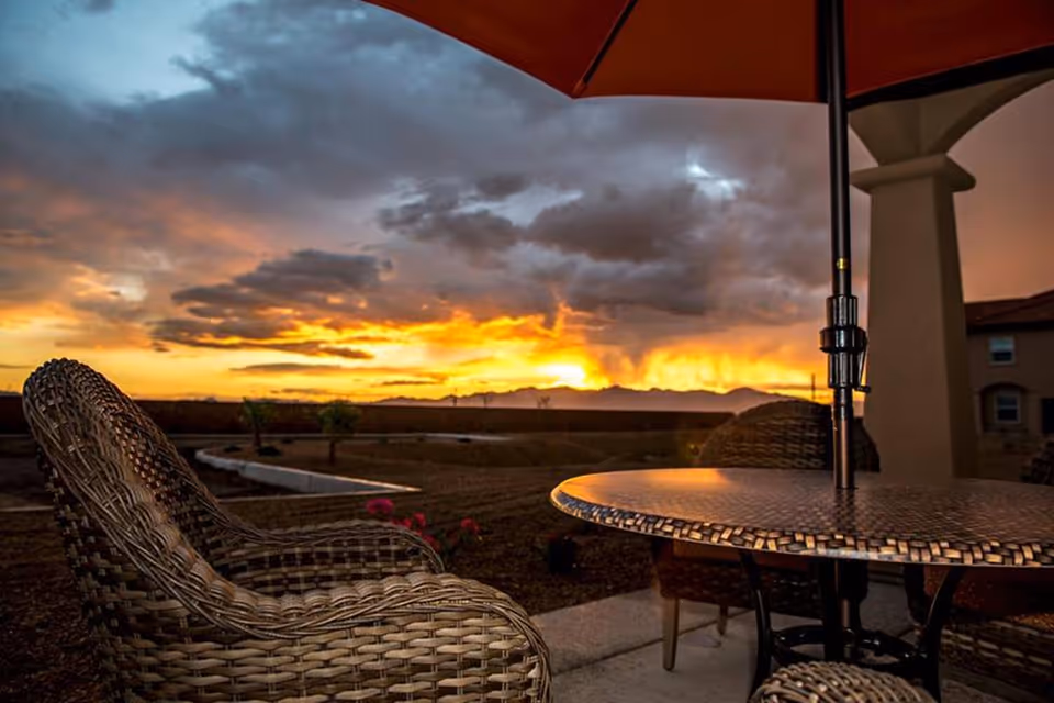 Wicker patio chairs and a round table with an umbrella on a patio overlooking a dramatic sunset and landscaped yard.