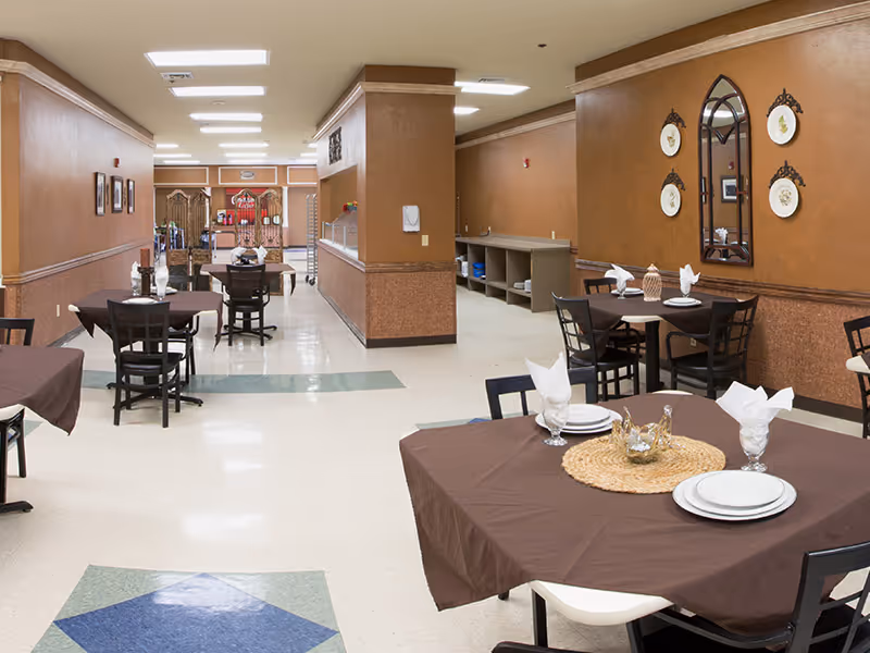 Dining room in a senior living facility with multiple tables set with brown tablecloths and place settings and a serving counter in the background.