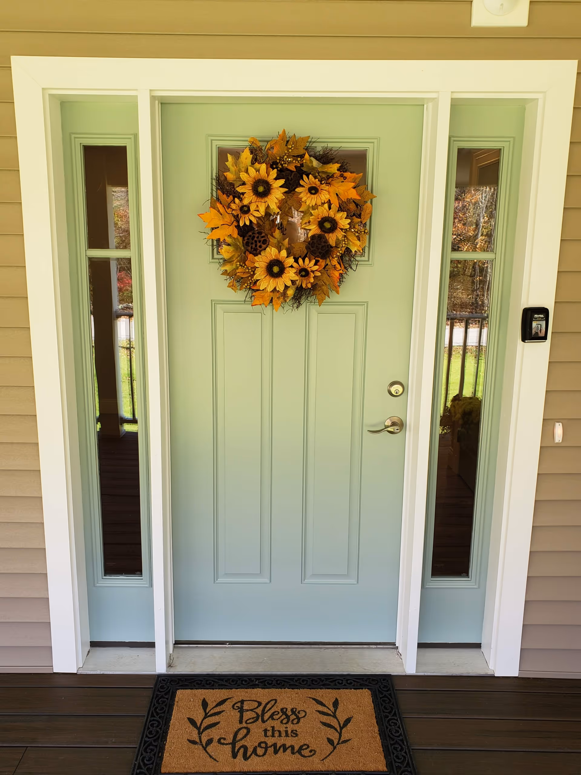 Front door of a house painted light green with a decorative wreath made of sunflowers and autumn leaves hanging on it. The door is flanked by two narrow vertical windows. A doormat with the words 'Bless this home' is placed in front of the door on a wooden porch.