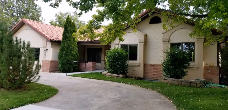 Exterior view of a single-story building with beige stucco walls and a red tile roof, surrounded by green trees and shrubs. A curved concrete pathway leads to the entrance, which has a small ramp and is partially shaded by a tree.