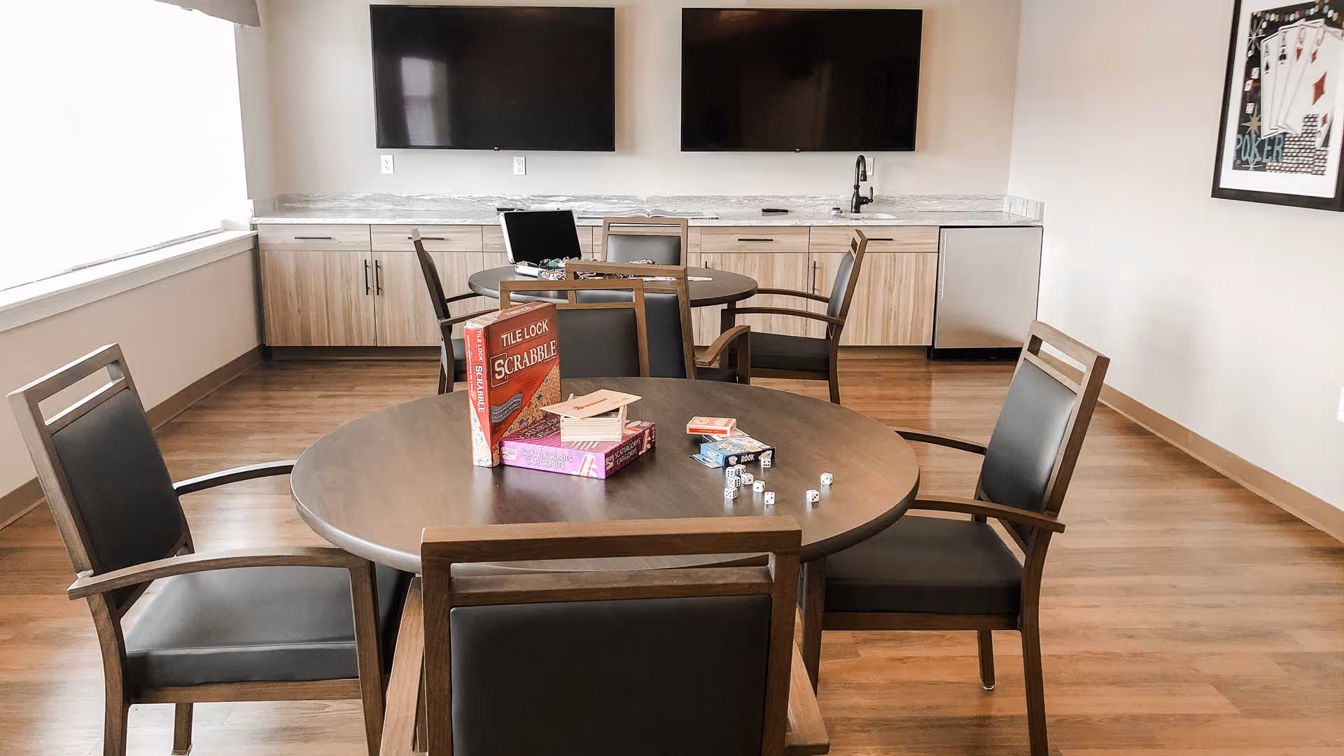 A common room with wooden floors and light-colored walls featuring two round tables with chairs around them. On the nearest table, there are board games including The Lock Scrabble, a deck of cards, and dice. The back wall has a countertop with cabinets, a sink, a small refrigerator, and two large flat-screen TVs mounted above.