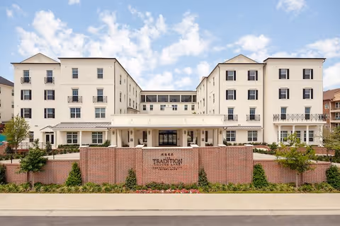 Front exterior view of The Tradition-Lovers Lane senior living facility, featuring a large multi-story white building with multiple windows and balconies, a brick wall with the facility's name and address, landscaped greenery, and a clear blue sky with scattered clouds.