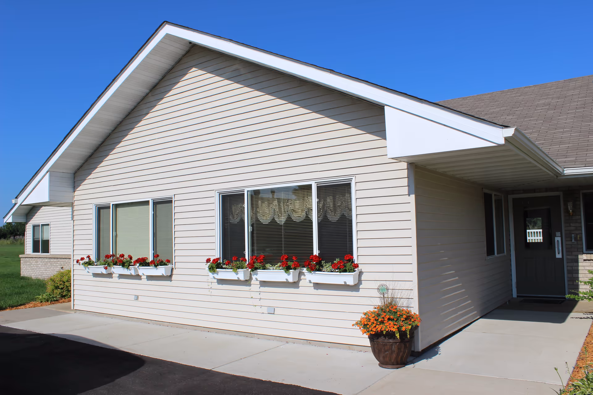 Exterior view of a beige single-story building with a sloped roof, featuring three windows with flower boxes filled with red flowers and a potted plant with orange flowers near the entrance door under a small covered porch.