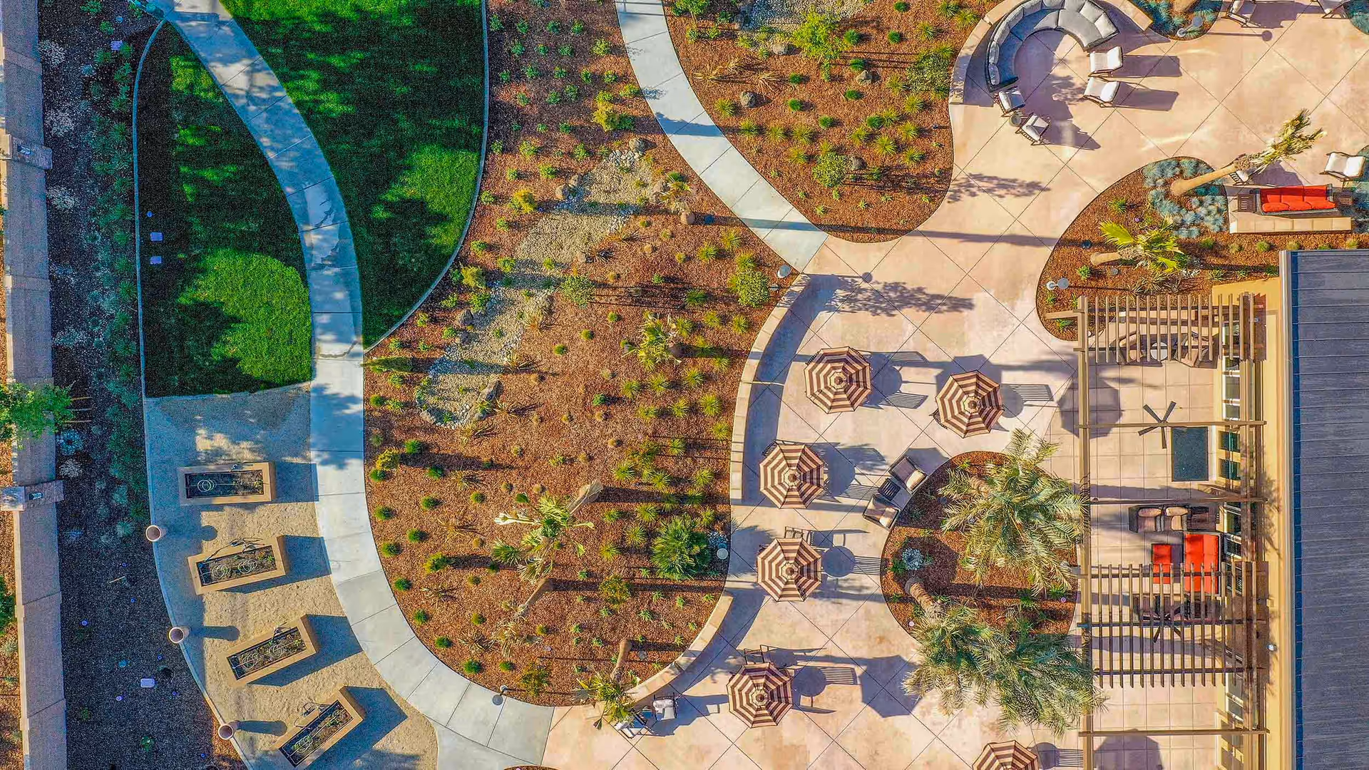 Aerial view of an outdoor patio area at Bruceville Point featuring multiple seating arrangements with umbrellas, curved walkways, landscaped garden beds, palm trees, and a pergola-covered seating area with red cushions.