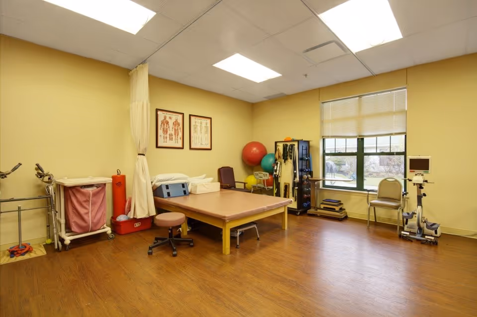 Therapy room with a treatment table, stool, exercise equipment, therapy balls, and anatomical charts on the wall.