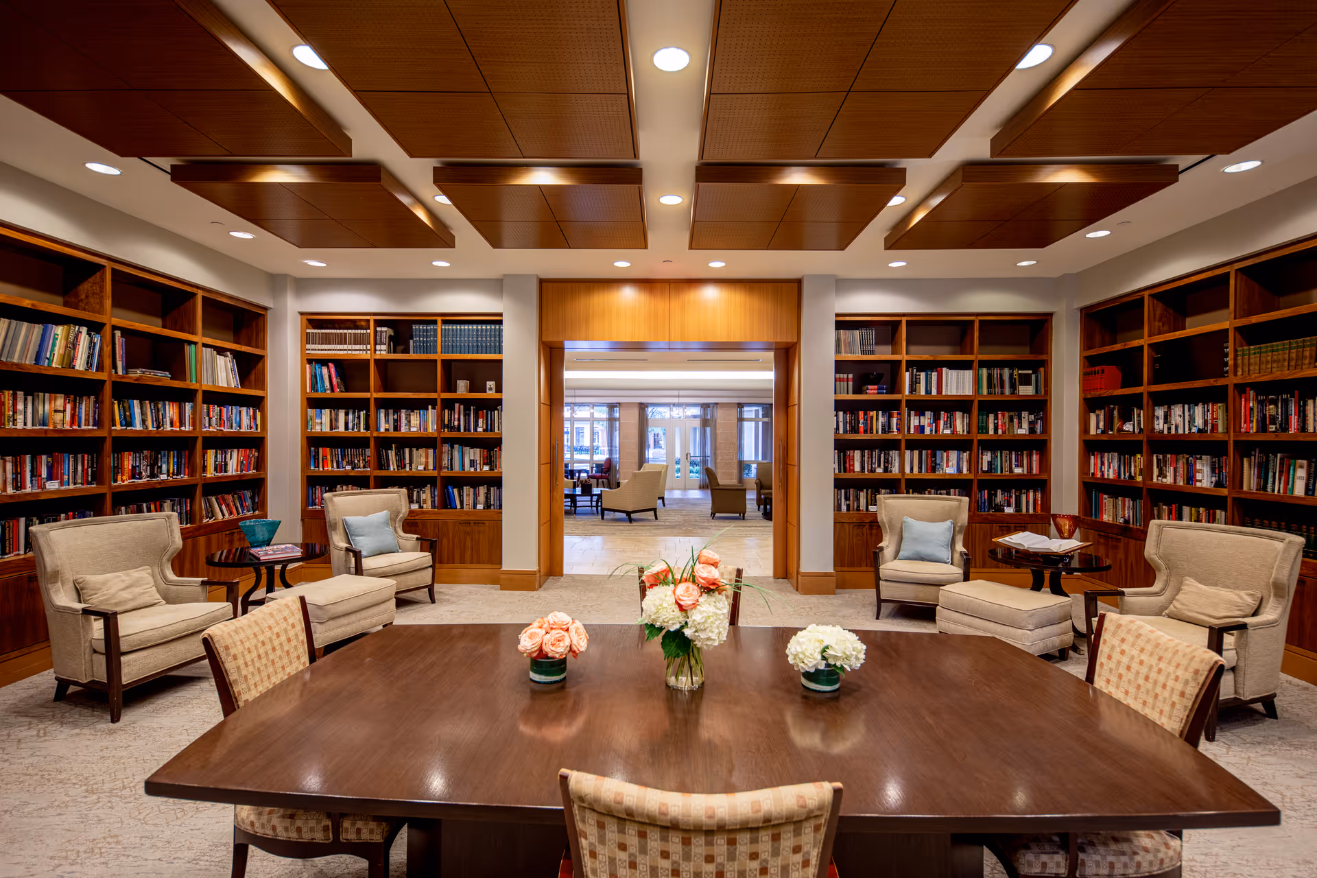 A cozy library room with wooden bookshelves filled with books lining the walls. There are four beige armchairs with cushions, two small round side tables, and a large wooden table in the center adorned with three floral arrangements. The ceiling features wooden panels with recessed lighting, and an open doorway leads to another seating area with more chairs and large windows.