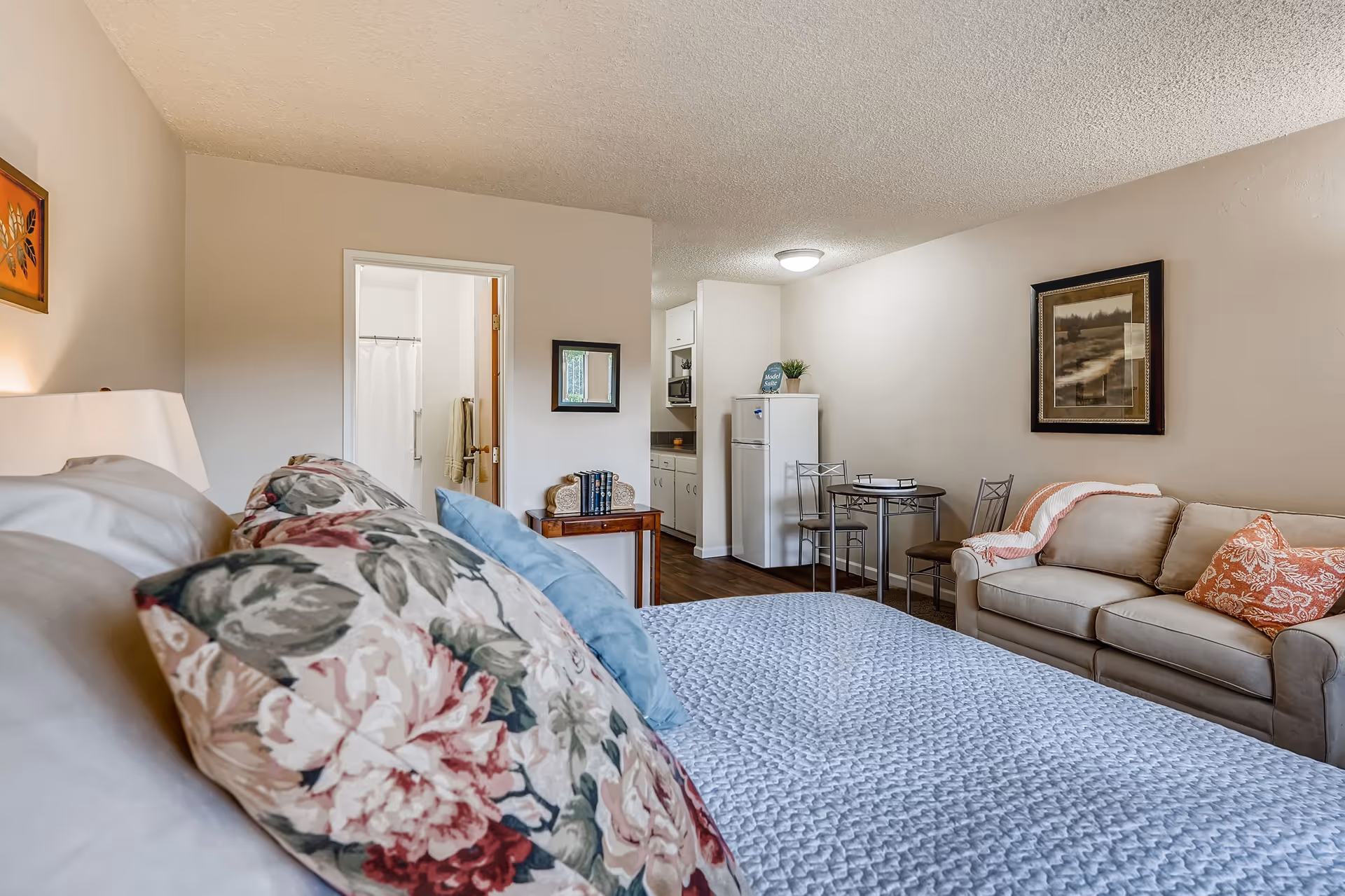 Interior view of a retirement facility room featuring a bed with floral pillows in the foreground, a small dining table with two chairs, a beige couch with decorative pillows and a throw blanket, a kitchenette with a refrigerator and cabinets, and a bathroom visible through an open door.