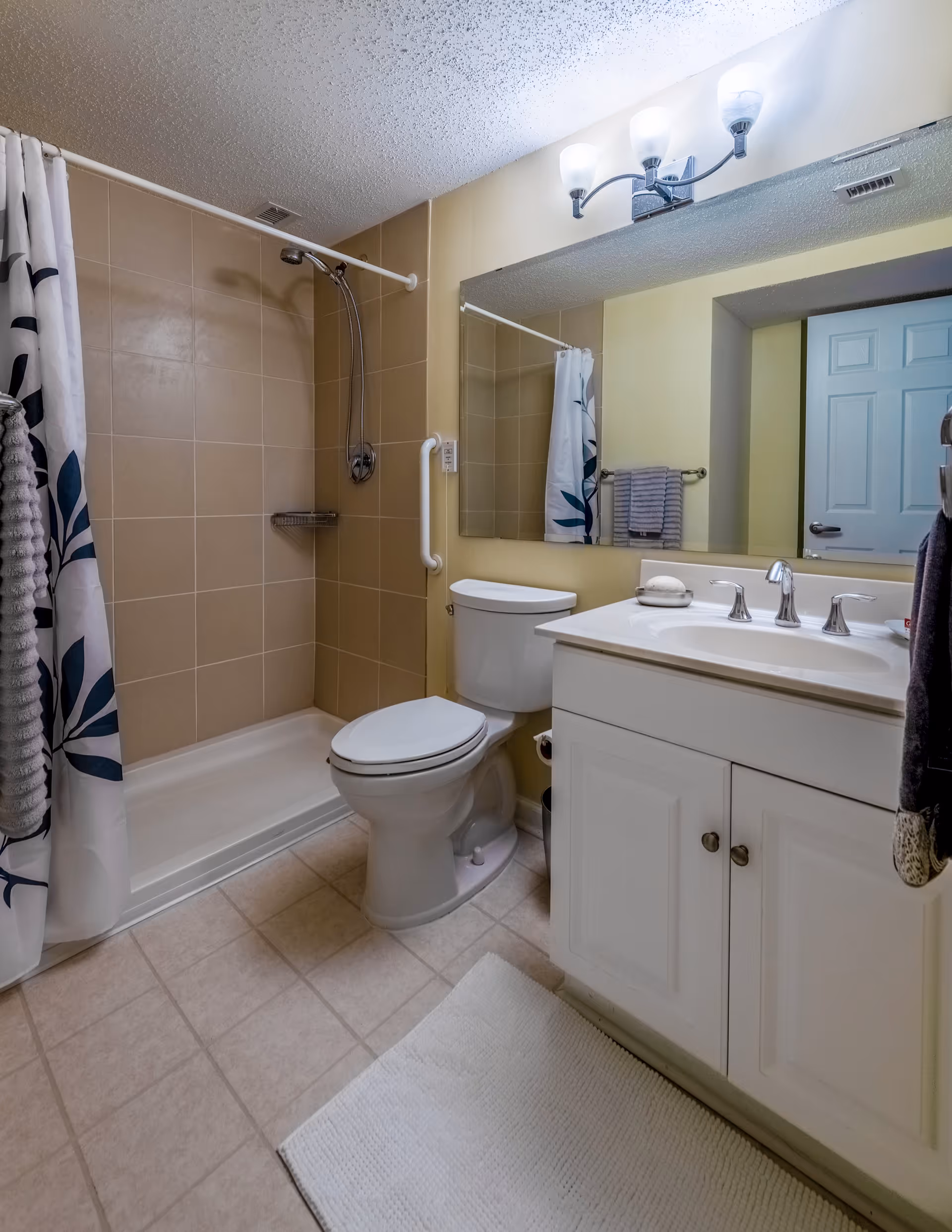 A clean bathroom featuring a walk-in shower with a white and blue floral shower curtain, beige tiled walls, a white toilet, a white vanity with a sink and silver faucet, a large mirror above the sink, a towel rack with a gray towel, and a white textured bath mat on the tiled floor.