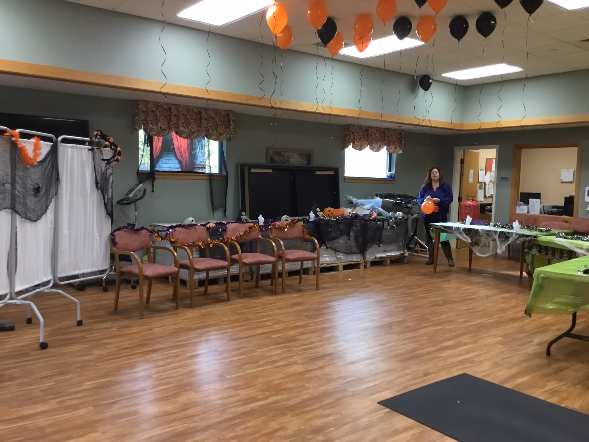 A decorated multipurpose activity room with chairs, tables, Halloween balloons and decorations and a person holding an orange balloon.