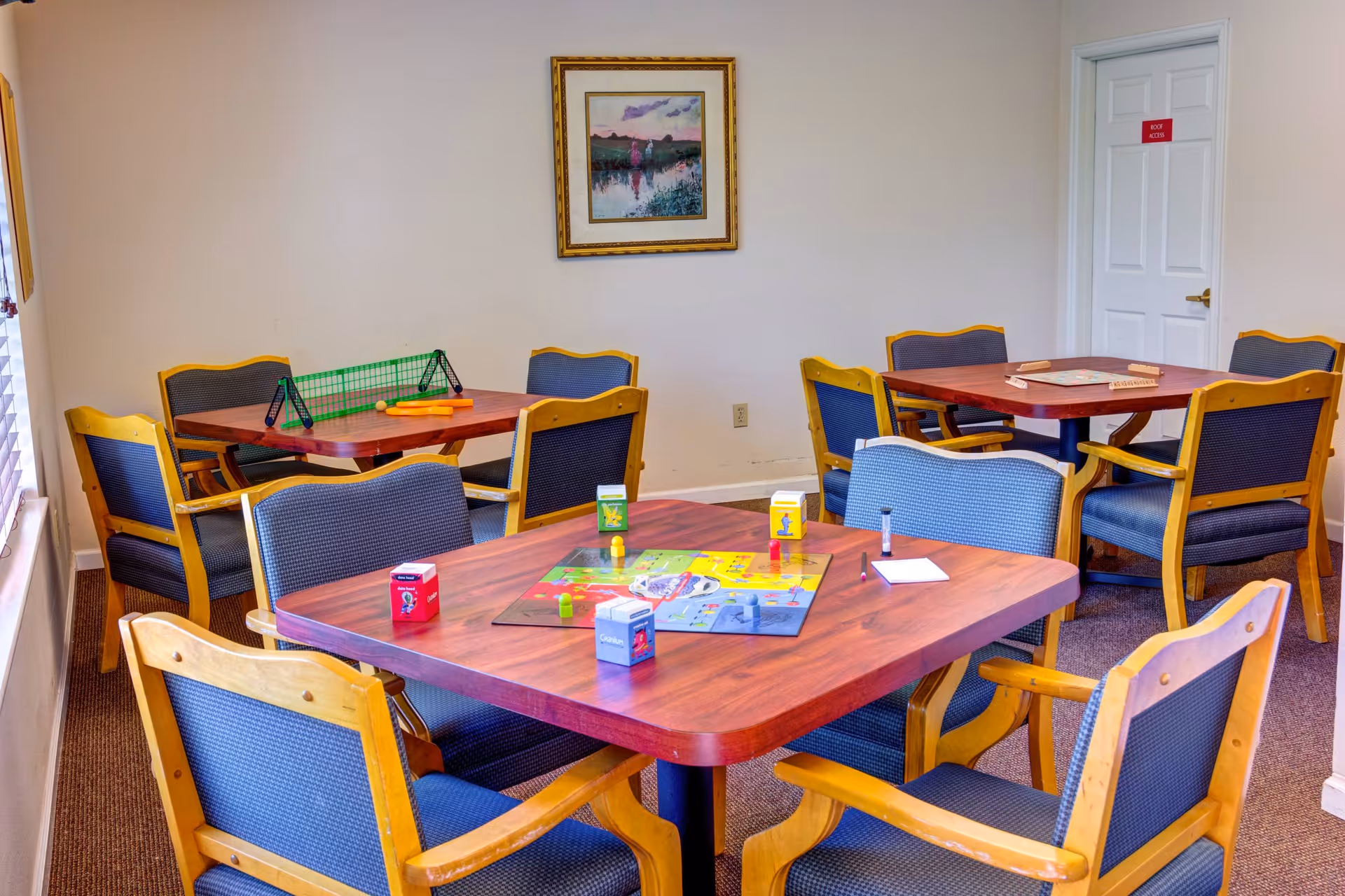 Community activity room with several wooden tables and upholstered chairs, a board game and game pieces set out on the nearest table.