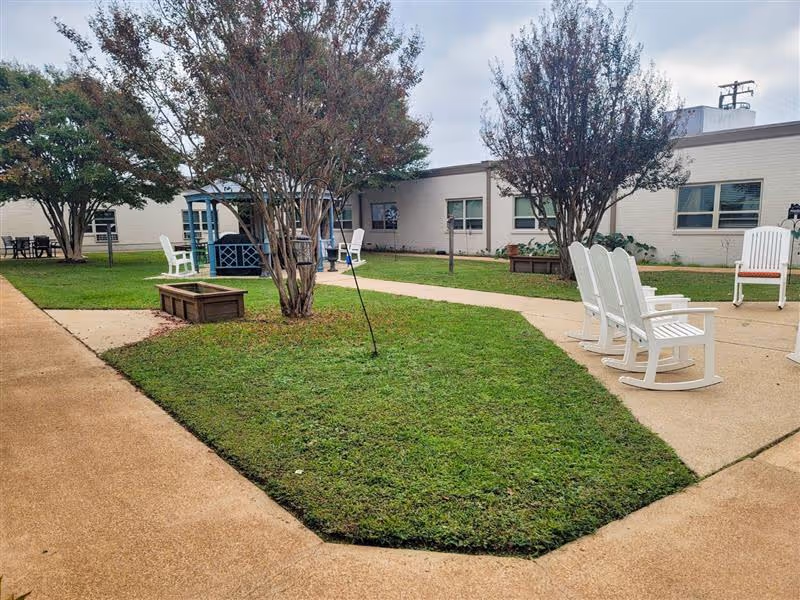 Outdoor courtyard area at Gilmer Nursing & Rehabilitation with green grass, several trees, white rocking chairs, a blue gazebo, and a beige paved walkway surrounding the grassy area. The building with white brick walls and windows surrounds the courtyard.