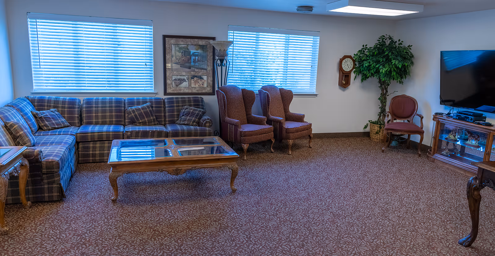 A senior living community lounge area with a plaid sectional sofa, two upholstered armchairs, a glass-top wooden coffee table, a wall clock, a potted plant, a flat-screen TV on a wooden stand, and two windows with blinds letting in natural light.