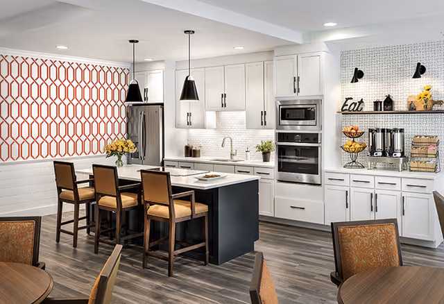 Modern kitchen with white cabinetry, stainless steel appliances, and a central island with four wooden chairs. The kitchen features a patterned red and white accent wall, pendant lights hanging over the island, and a countertop area with coffee and snack station labeled 'Eat'. The floor is wood-style, and there are round tables with chairs in the foreground.