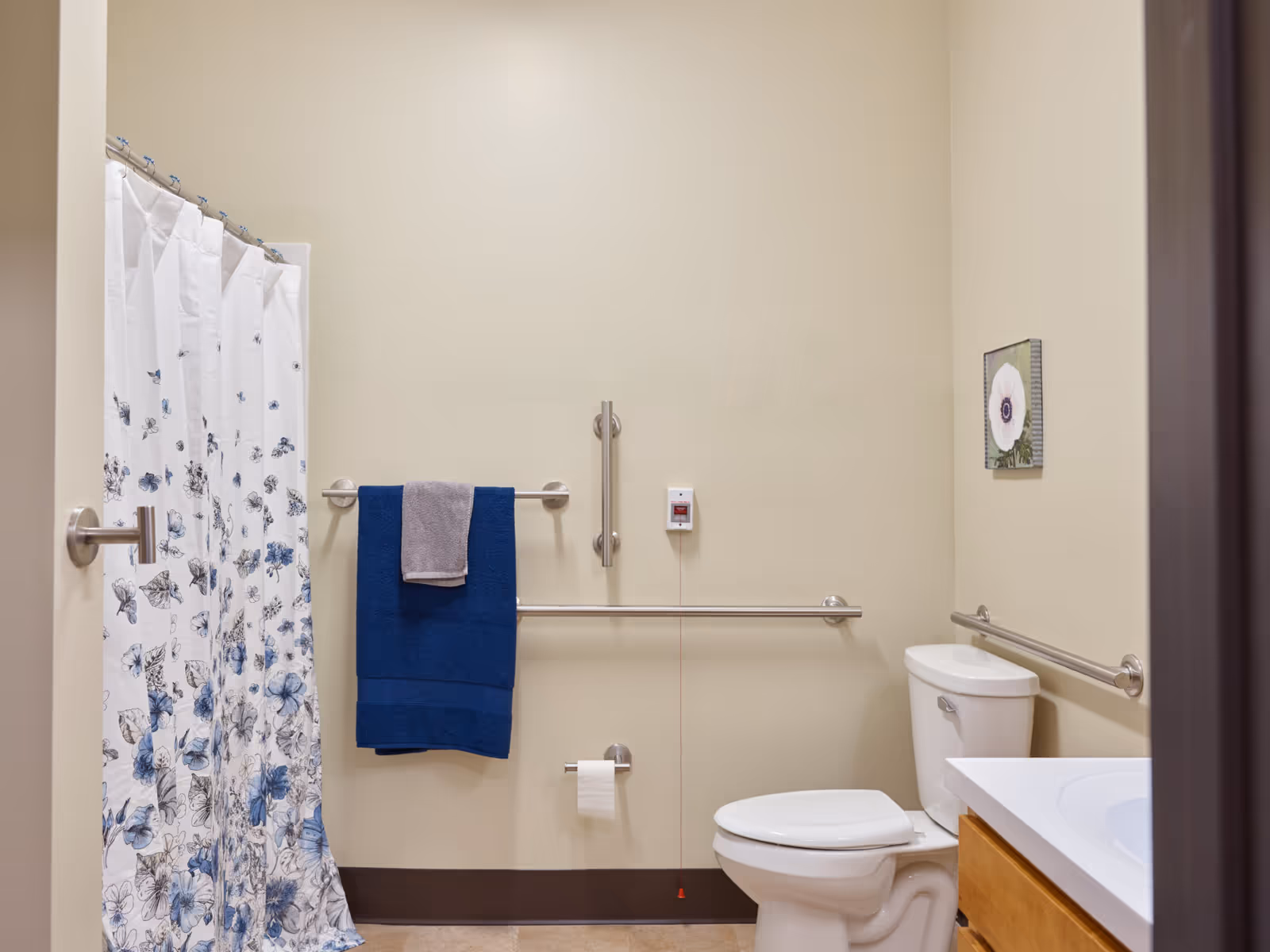 A bathroom with beige walls featuring a white toilet, a white sink with a wooden cabinet, and a shower with a white curtain decorated with blue and gray floral patterns. There are stainless steel grab bars on the walls, a towel rack with a blue and gray towel, a toilet paper holder, and a small framed flower picture on the wall.