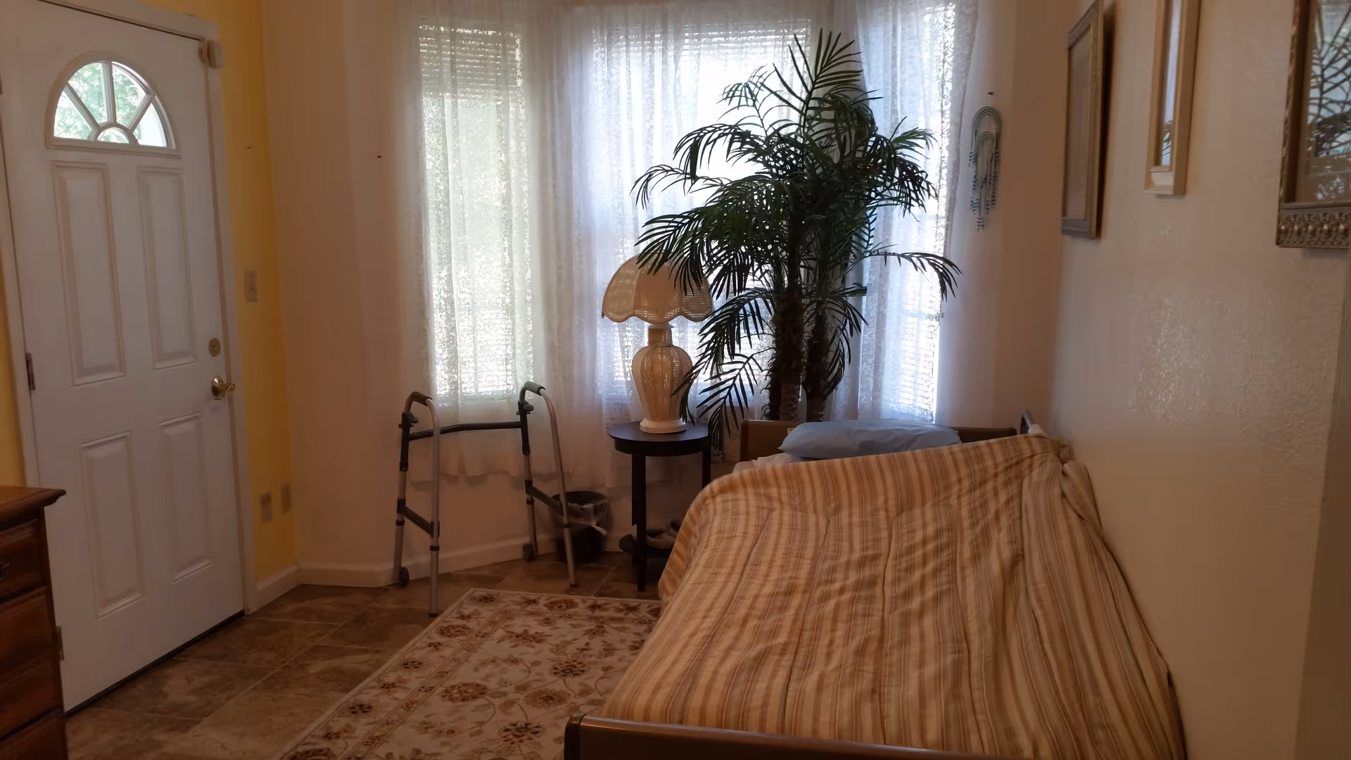 A small living area with a striped couch, a side table with a lamp, a large potted plant, a walker, and a white door with a window. The room has tiled flooring and sheer curtains covering the windows.