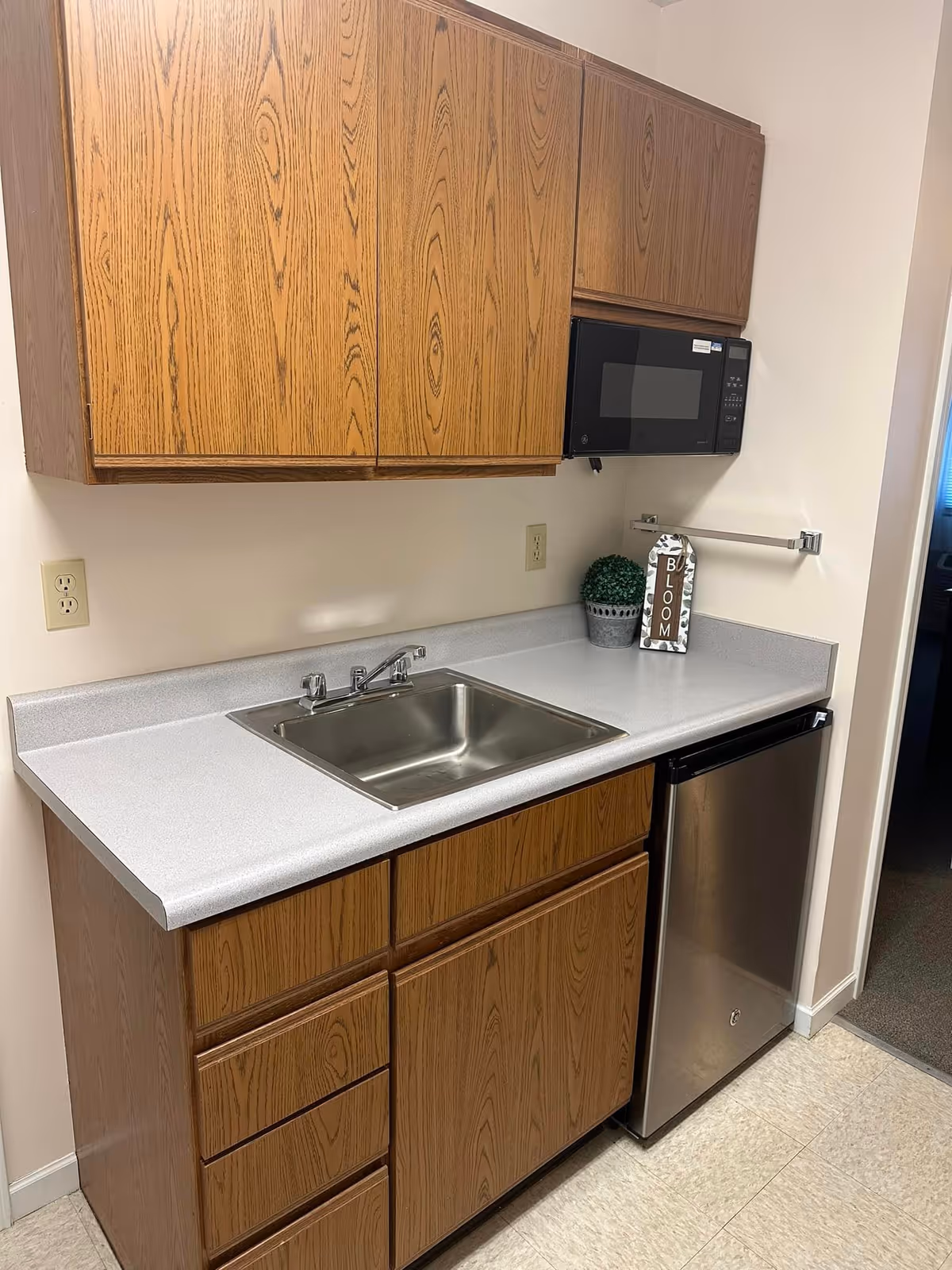 A small kitchenette area with wooden cabinets above and below a white countertop. The countertop has a stainless steel sink with a faucet. Above the counter is a black microwave mounted under the upper cabinets. To the right of the sink is a small stainless steel refrigerator. On the countertop, there is a small potted plant and a decorative sign that says 'BLOOM'. The walls are light-colored, and the floor has light-colored tiles.