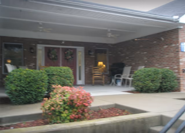 Covered entrance area of a brick building with double doors decorated with wreaths, surrounded by green bushes and a flowering shrub in front. There are chairs and a lamp visible on the porch.