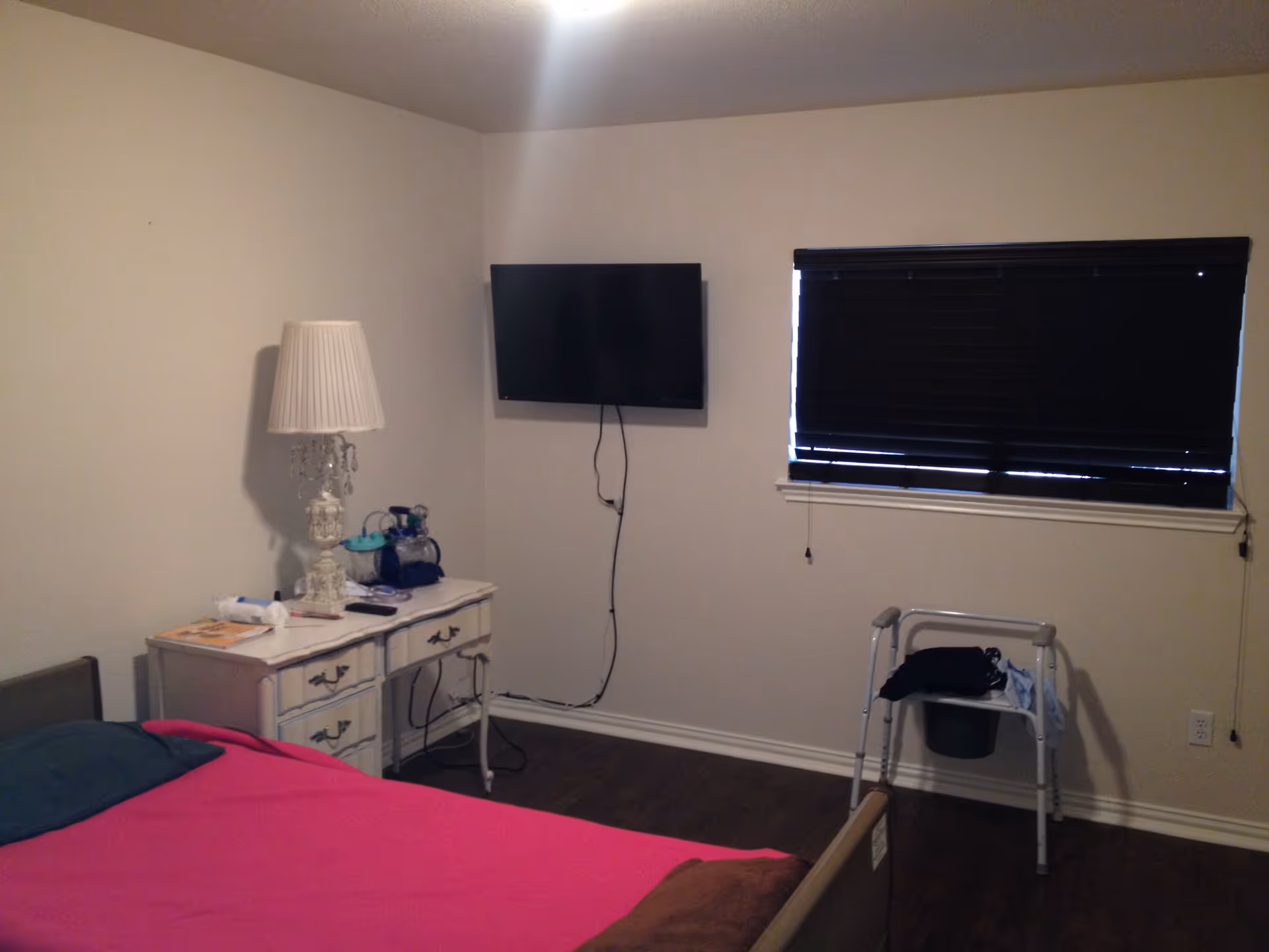 A bedroom with a bed covered in a bright pink blanket and a teal pillow. Next to the bed is a white vintage-style nightstand with a decorative lamp and some items on top. A flat-screen TV is mounted on the wall above the nightstand. There is a window with closed black blinds and a walker with some clothes draped over it near the window. The floor is dark wood and the walls are light-colored.