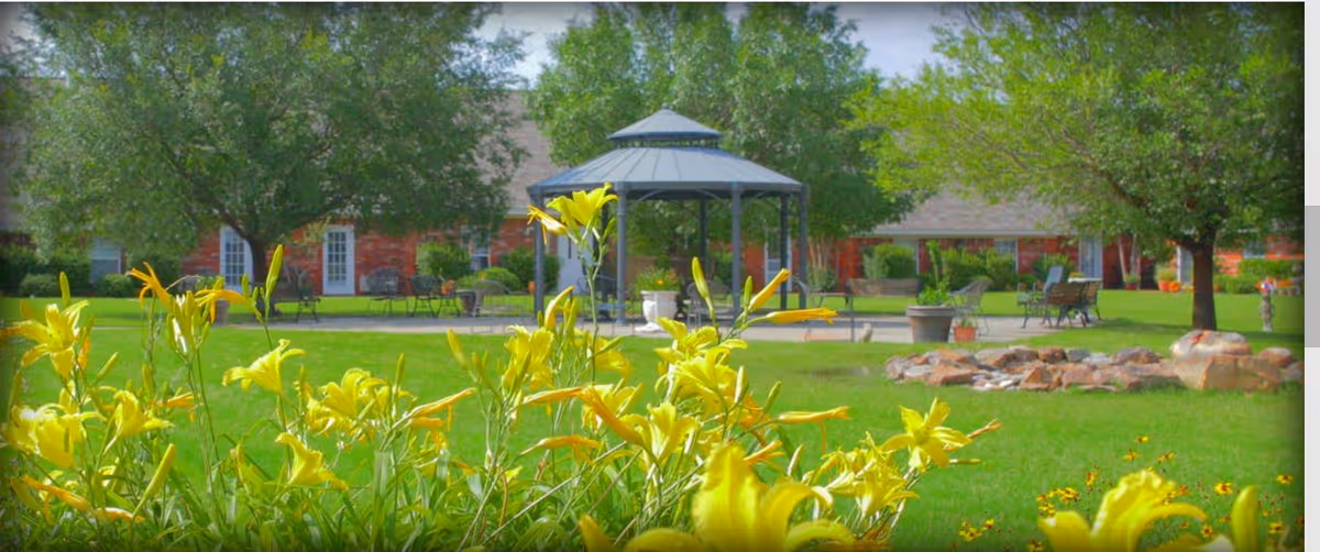 A bright outdoor garden area at Brookridge Retirement Community featuring yellow flowers in the foreground, a green lawn, trees, a gazebo, benches, and a fire pit with rocks.