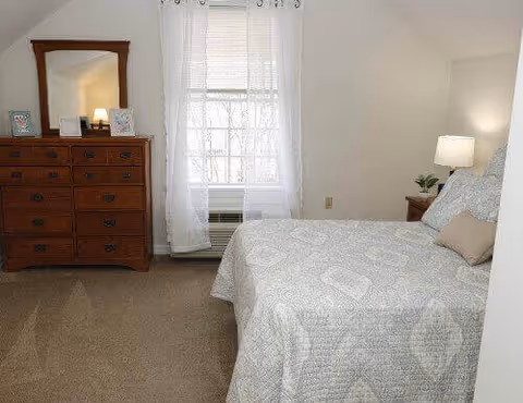 A cozy bedroom with a bed covered in patterned bedding, a nightstand with a lamp, a dresser with mirror, and a window with sheer curtains.