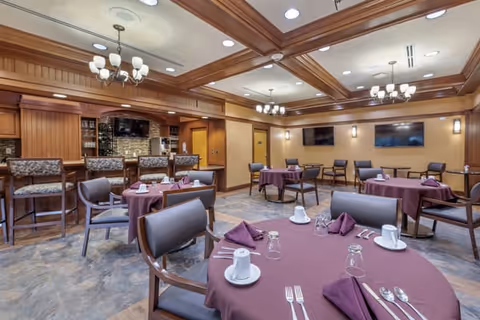 A warm dining room with round tables draped in purple tablecloths, place settings and cushioned chairs, chandeliers, and a wood-paneled bar area.