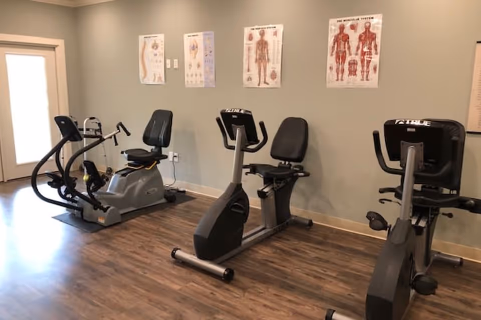 A small exercise room with three stationary recumbent bikes on a wooden floor. The walls are light-colored and have anatomical posters showing human muscles and body parts. A door with a window is visible on the left side.