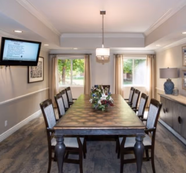 A well-lit dining room with a long wooden table surrounded by eight chairs. A floral centerpiece decorates the middle of the table. Two large windows with beige curtains allow natural light to enter the room. A modern light fixture hangs above the table. On the right side, there is a sideboard with a blue lamp and decorative items. A flat-screen TV is mounted on the left wall displaying text.