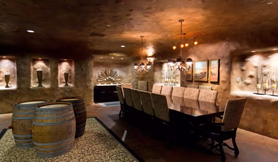 A dimly lit dining room with a long dark wooden table surrounded by upholstered chairs. The walls have a textured, rustic finish with built-in niches displaying decorative vases and candles. Three wooden barrels are placed on a patterned rug near the table. Two chandeliers with warm lighting hang from the ceiling, and framed artwork is displayed on one wall.