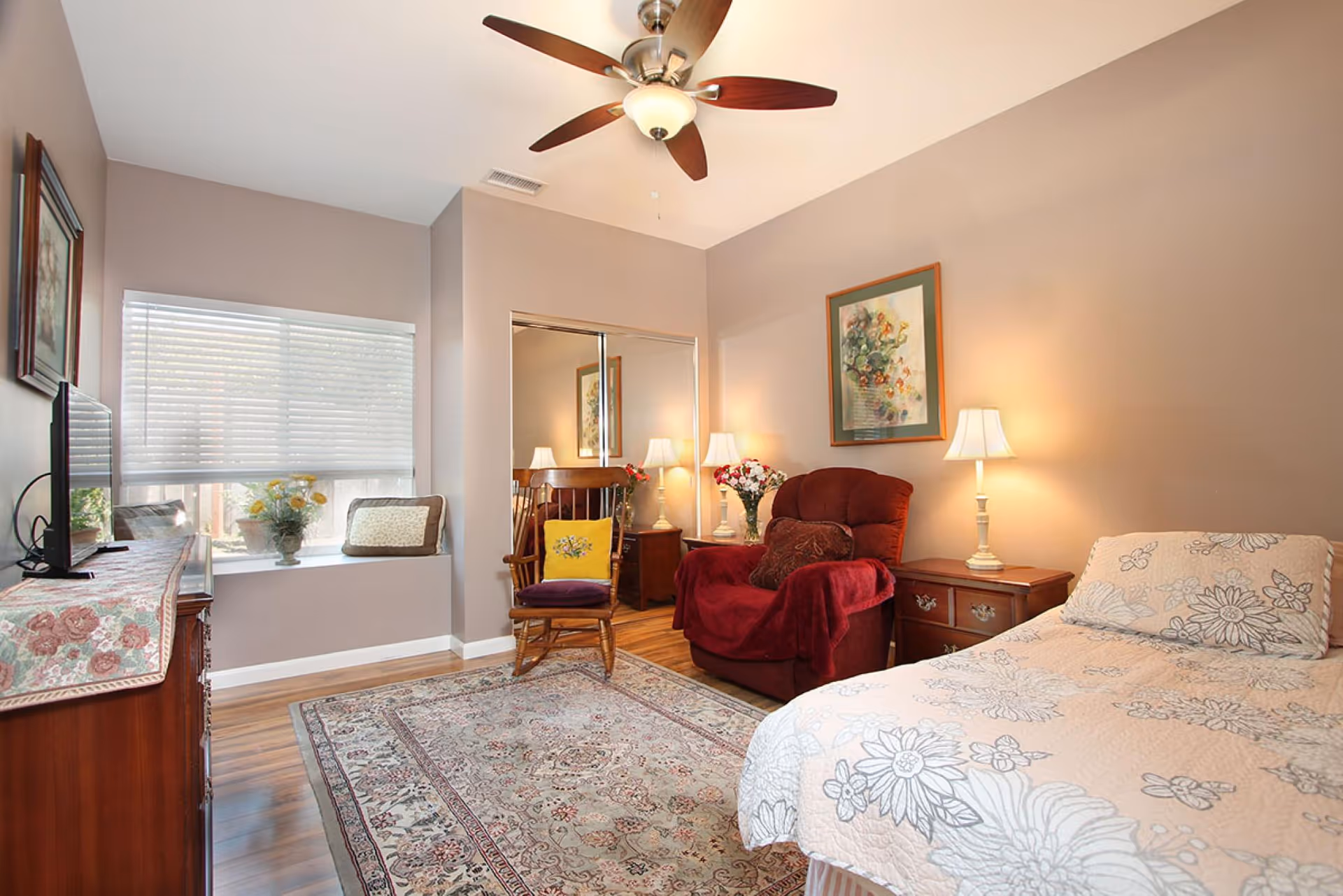 A cozy bedroom with a single bed covered in a floral bedspread, a red armchair with a pillow and a throw blanket, a wooden rocking chair with a yellow cushion, two bedside tables with lamps, a large window with blinds, a ceiling fan with lights, a framed floral painting on the wall, a wooden dresser with a TV on top, and a patterned area rug on the wooden floor.