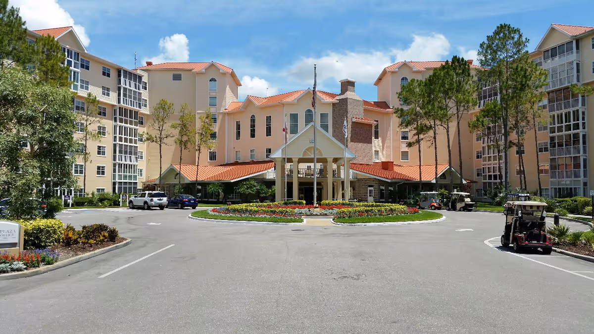 Front exterior view of Freedom Plaza senior living facility with a circular driveway, landscaped flower beds, multiple flagpoles, and several golf carts parked near the entrance. The building has beige walls, red-tiled roofs, and multiple floors with windows and screened balconies.