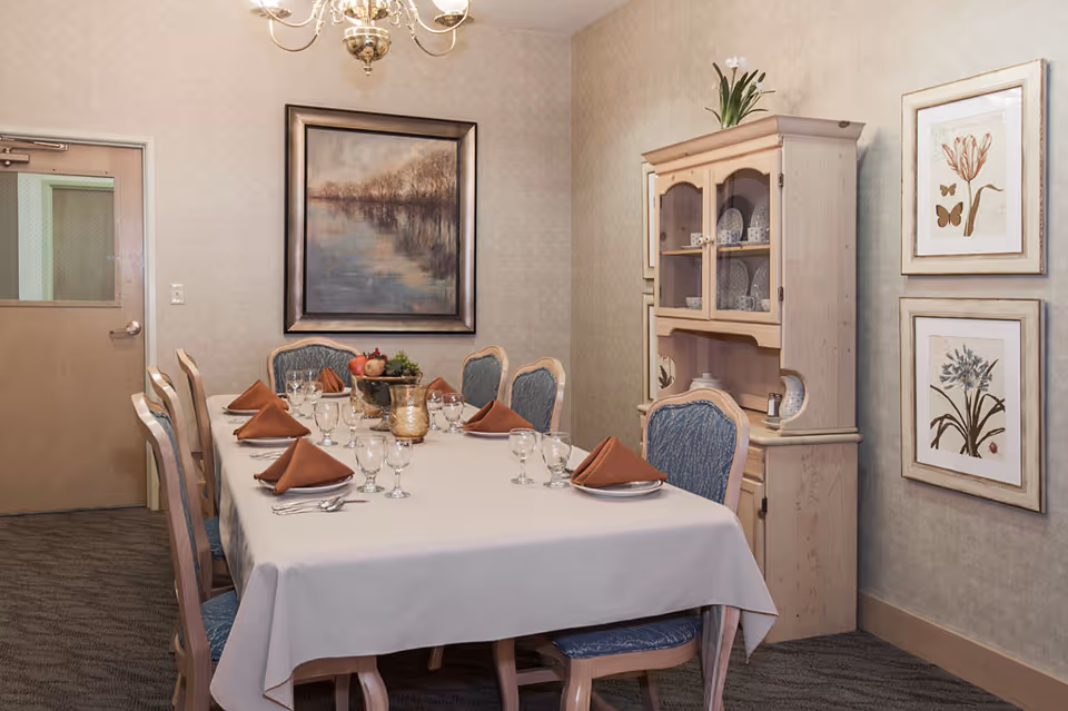 A dining room with a rectangular table covered with a white tablecloth, set with plates, glasses, silverware, and brown folded napkins. There are eight upholstered chairs around the table. On the right side, there is a wooden cabinet with glass doors displaying dishes. The walls are decorated with framed paintings of flowers and a landscape. A chandelier hangs from the ceiling.